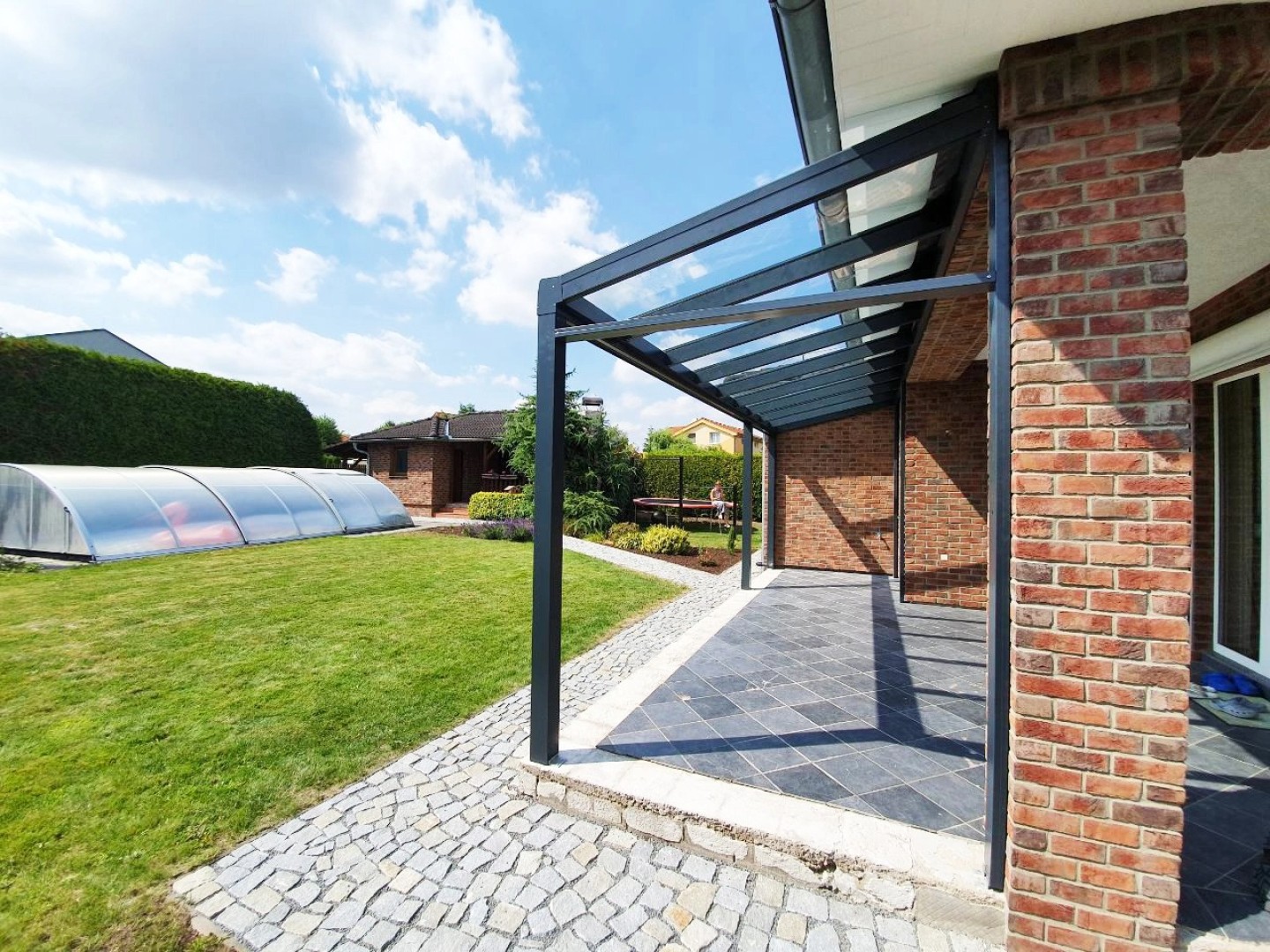 Moderní antracitová hliníková pergola se skleněnou střechou u cihlového domu se zahradou a bazénem.